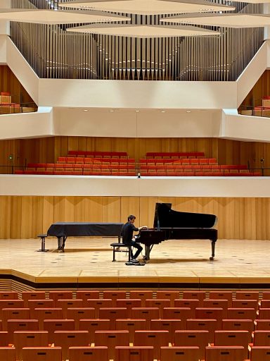 Ein Musiker spielt Klavier auf einer leeren Konzertbühne mit Holzverkleidung.