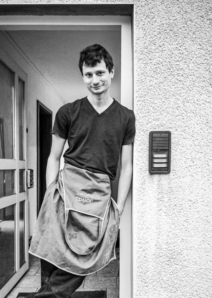 Junger Mann in T-Shirt und Schürze steht entspannt vor einer Tür.